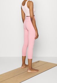 Ljusrosa atletisk crop top i kombination med matchande capri leggings, med en slät yta och en figurnära design. Står på en yogamatta.