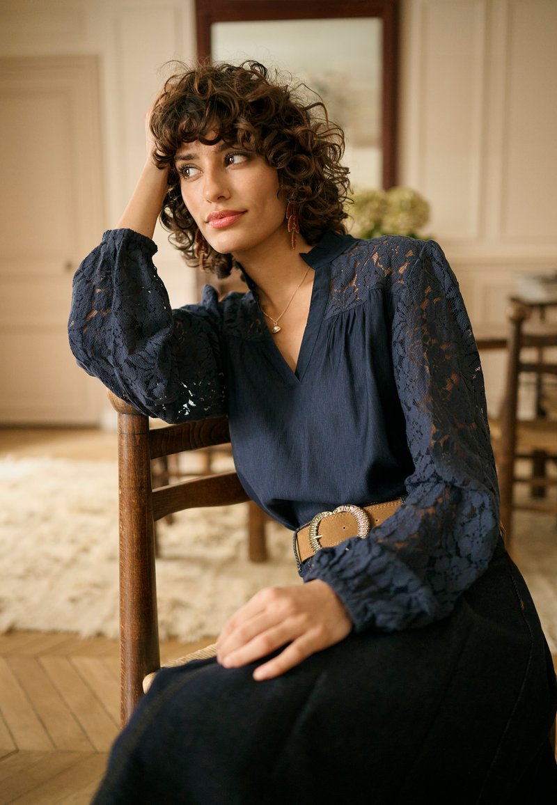 Blouse bleu marine avec manches en dentelle, encolure en V et tissu texturé. Stylisée avec une ceinture marron ornée d'une boucle argentée décorative.