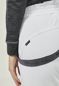 Weiße dehnbare Hosen mit einem elastischen Bund, ausgestattet mit einem dunkelgrauen verstellbaren Gürtel und einem schwarzen Textillabel nahe der hinteren Naht.