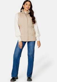Béžový péřový vesta s vysokým límcem a stahovací šňůrkou v pase, nošená přes krémový svetr a v kombinaci s modrými džíny a černými botami.