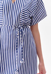 Sukienka z paskowanej tkaniny w niebieskie i białe pionowe paski, z dekoltem w kształcie litery V i wiązanym w pasie. Krótkie rękawy i miękka faktura.
