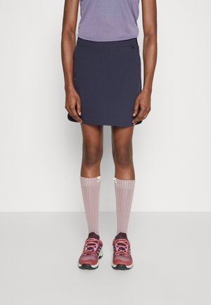 Osoba ubrana w fioletową bluzkę, granatową spódnicę za kolano, różowe podkolanówki i różowe trampki, stojąca na tle gładkiej białej ściany.