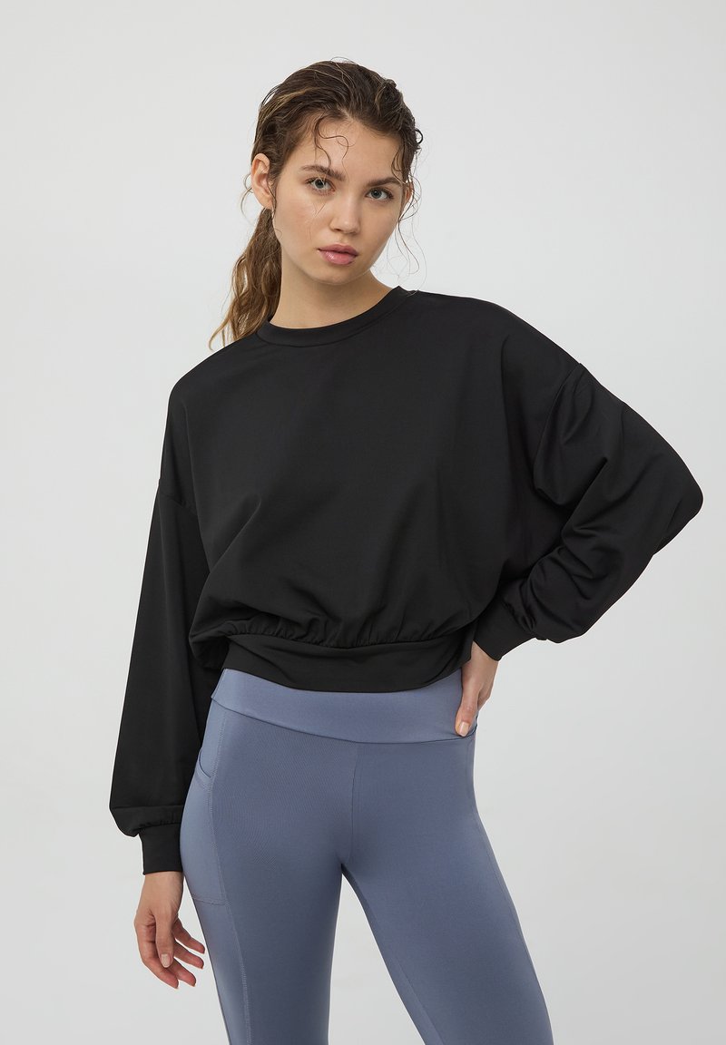 Czarny skrócony sweter z luźnymi rękawami, elastycznym paskiem w talii, zestawiony z jasnoniebieskimi legginsami o wysokim stanie. Gładki materiał, minimalistyczny design.