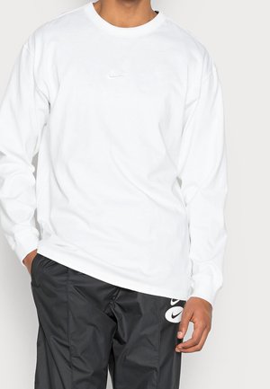 Mężczyzna ubrany w białą, długorękawową koszulkę Nike i czarne spodnie Nike z logo, stojący z jedną ręką w kieszeni na jednolitym tle.