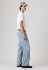 Νεαρός άνδρας που φοράει ανοιχτό μπλε φαρδιά τζιν παντελόνια, λευκό μπλουζάκι, γκρι καπέλο και μαύρα παπούτσια, στέκεται πλευρικά μπροστά σε ένα απλό ανοιχτό φόντο.