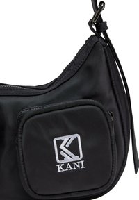 Svart stoff skulderveske med glidelås hovedrom, frontlomme og brodert "KANI"-logg. Glatt tekstur med justerbar stropp.