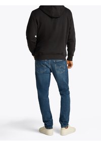 Felpa nera con cappuccio e jeans blu. I jeans presentano tasche posteriori con dettagli di cucitura. Le sneaker sono beige, con una texture liscia.