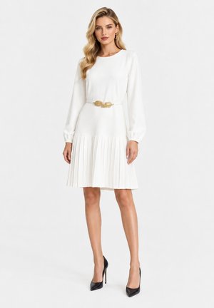 Donna che indossa un vestito bianco a maniche lunghe con gonna plissettata, cintura dorata a foglia e scarpe nere con tacco alto, in piedi davanti a uno sfondo semplice.