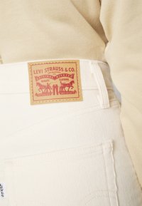 Gros plan sur un pantalon en velours côtelé blanc Levi's montrant une étiquette en cuir Levi Strauss & Co. au-dessus de la poche arrière et un haut beige.