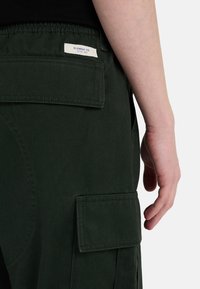Groene cargobroek van een textuurstof, met een achterzak met klep en een klein label met de tekst "ELEMENT CO." op de tailleband.