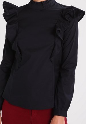 Czarna bluzka z długim rękawem, falbanami na ramionach i wysokim kołnierzem, zestawiona z czerwonymi spodniami.