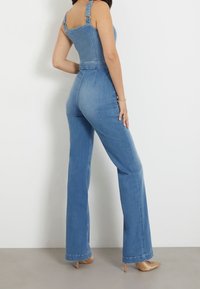 Une salopette en denim bleu clair avec un corsage ajusté, des jambes larges, des bretelles ajustables et des détails de couture visibles.