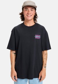 Camiseta de algodón negra con mangas cortas, que presenta un colorido logo gráfico en el lado izquierdo del pecho. Ajuste relajado y diseño minimalista destacados.