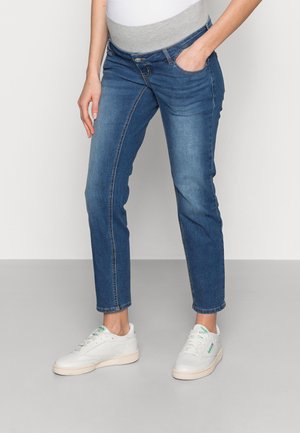 Person iført blå graviditetsjeans med grå elastisk talje og hvide sneakers, stående med den ene hånd i lommen.