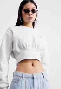 Lys grå kort sweatshirt med samlet midje, lange ermer, og en liten logo på brystet, tilpasset lys blå høytlivsjeans.