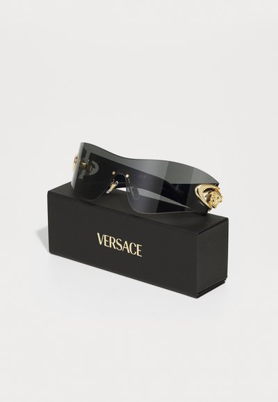 Svarta överdimensionerade solglasögon med guldfärgad detaljer, med lejonmotiv på bågarna, vilande på en svart Versace-låda.