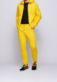 Gelbfarbener Hoodie und Jogginghose aus weichem Material, mit einem Frontreißverschluss und schwarzen Akzenten. Kombiniert mit schwarzen und gelben Sneakern.