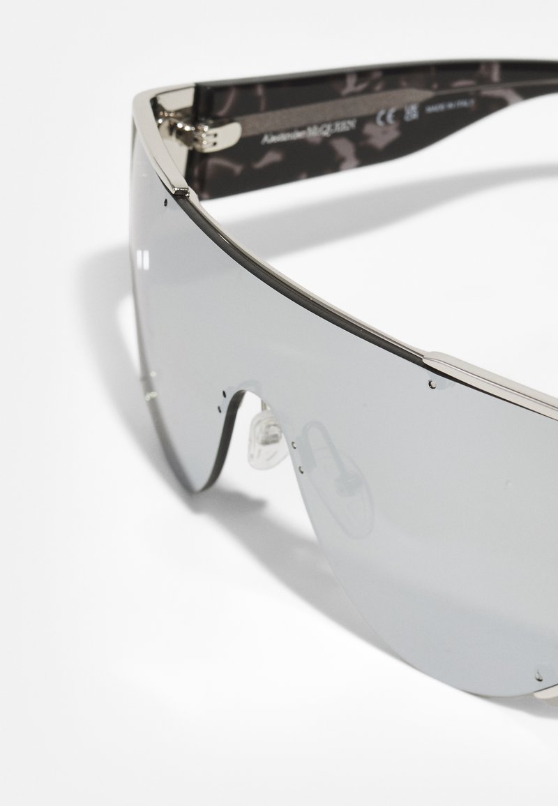 Sonnenbrille mit großen, dunkelgrauen Gläsern, silbernem Rahmen und transparenten Nasenpads. Gemusterte Details an den Bügeln, versehen mit dem Markenlogo.