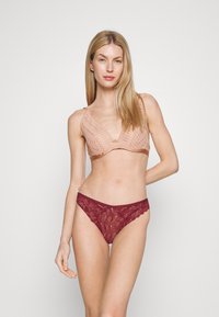 Koronkowy bralet w delikatnym odcieniu brzoskwini, z dekoltem w kształcie litery V i regulowanymi ramiączkami. W zestawie przezroczysta koronkowa stringi w kolorze bordowym z falbaniastymi brzegami.