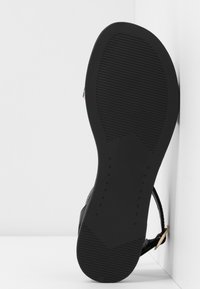 Semelle de sandale noire avec motif de grip texturé, affichée verticalement sur un fond blanc en coin, montrant la semelle extérieure et la bride de cheville.