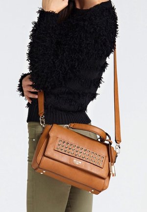 Femme portant un pull noir texturé et un pantalon olive, portant un sac à main marron Guess avec un rabat avant clouté et une bandoulière ajustable.