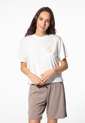 Frau mit dunklen Haaren, die ein weißes T-Shirt mit gelbem Text "Soup of the Day" und weiten beigen Shorts trägt, steht vor einem schlichten weißen Hintergrund.