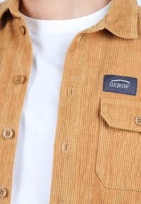 Chemise en velours côtelé beige avec un design à boutons, dotée d'une poche poitrine, d'un label OXBOW et de boutons en bois, portée sur un t-shirt blanc.