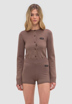 Kvinde iført en brun knappet cardigan og matchende strikkede shorts med høj talje med sorte mærkepatche på bryst og shorts.