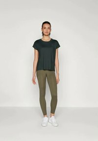 Mørkegrønn t-skjorte med korte ermer, kombinert med olivengrønne leggings. Modellen bruker hvite sneakers. Glatt, lett stoff med en avslappet passform.