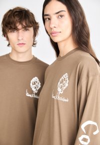 Brown long-sleeve t-shirts feature white artichoke graphics and text on the front and sleeve, made from soft fabric with a relaxed fit.