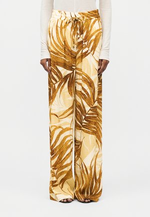 Hlače širokih nog z rjavim in bež tropskim vzorcem listja, z vrvico v pasu, kombinirane z dolgim kremnim zgornjim delom in črnimi sandali z odprtimi prsti.