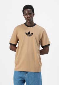 Šviesiai rudos trumpomis rankovėmis marškiniai su juodu apykakle ir rankovių kraštais, kurių priekyje išsiuvinėtas juodas Adidas logotipas. Pagaminta iš minkštos medvilnės medžiagos.