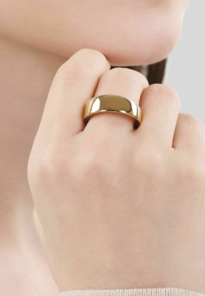 Primer plano de una mano cerca del mentón que lleva un anillo dorado liso, ancho y brillante en el dedo medio sobre un fondo neutro.