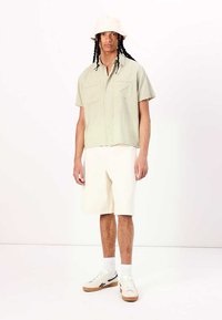 Camicetta verde chiaro a maniche corte, con bottoni e due tasche, abbinata a pantaloni corti bianchi fino al ginocchio e un cappello a secchiello beige. Sneakers con suola in gomma.