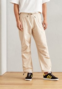 Beige, leichte Hose mit elastischem Bund und Kordelzug. Dazu schwarze Sneakers mit gelben Streifen.