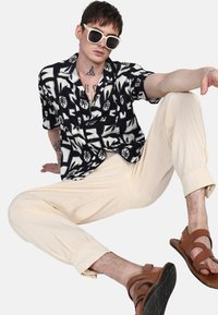 Camicia a righe con motivo di foglie in bianco e nero, leggera, con bottoni; abbinata a pantaloni beige plissettati e sandali aperti marroni.