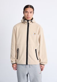 Chaqueta de forro polar beige con cierre de cremallera negra, capucha y bolsillos laterales. Incluye un pequeño logotipo en el pecho y detalles de cordón en la capucha.
