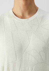 Gros plan sur une personne portant un pull en tricot crème avec un motif géométrique texturé et un col rond.