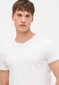 Weißes Baumwoll-T-Shirt mit kurzen Ärmeln und einem runden Halsausschnitt, mit einem kleinen grauen Markenlogo im Bereich der linken Brust.