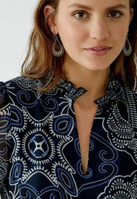 Marineblaue Bluse mit floralem Muster in Weiß und Blau, mit rüschenförmigem Kragen und V-Ausschnitt. Das Modell trägt silberne Ohrringe.