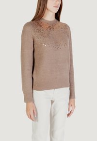 Maglione marrone con un design testurizzato e scintillante nella parte superiore. Maglia leggera, colletto a girocollo e polsini a costine. Indossato con pantaloni bianchi.