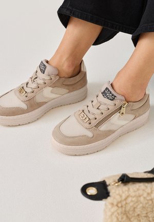Zapatillas beige con una mezcla de texturas lisas y de ante, que presentan una cremallera, un cierre de cordones en la parte delantera y una lengüeta estampada, sobre un fondo blanco.
