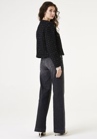 Zwarte tweed korte jas met een gestructureerd patroon, ronde halslijn, gecombineerd met high-waisted donkerblauwe jeans en lichtgekleurde hakken.