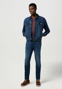 Denimjack met twee borstzakken en knoopsluiting, gedragen over een kastanjebruine blouse. Slim-fit donkerblauwe jeans en bruine veterschoenen.
