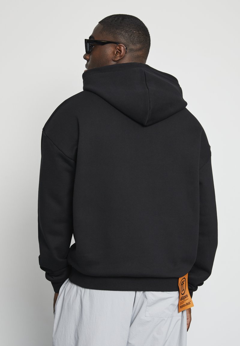 Black hoodie with a large hood, made of smooth fabric. Features an orange label at the hem and relaxed fit. Model wears sunglasses.
