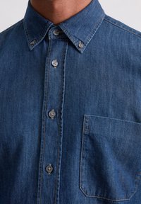 Chemise en denim bleu foncé, avec un col button-down, des boutons gris et une poche poitrine. Le tissu a une texture lisse et des coutures subtiles.