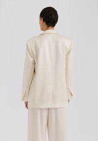 Krémovo sako so štruktúrovaným dizajnom, výrezovými klopami a jedným rozparkom. Hladká, saténová textúra s dlhými rukávmi.