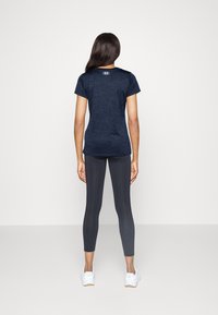 Kvinna som står vänd bort med en marinblå sportt-shirt, mörkgrå perforerade leggings och vita sneakers på en enfärgad bakgrund.