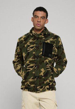 Mężczyzna ubrany w zieloną i brązową polarową kurtkę kamuflażową z czarną kieszenią na piersi zapinaną na zamek oraz beżowe spodnie cargo, stojący na tle szarego tła.