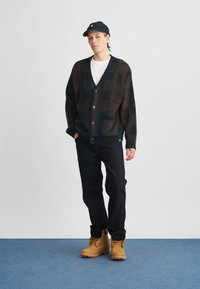 Hnedý a čierny károvaný kardigán s výstrihom do V, zapínaním na gombíky a vreckami, skombinovaný s čiernymi nohavicami a svetlohnedými topánkami, na jednoduchom pozadí.